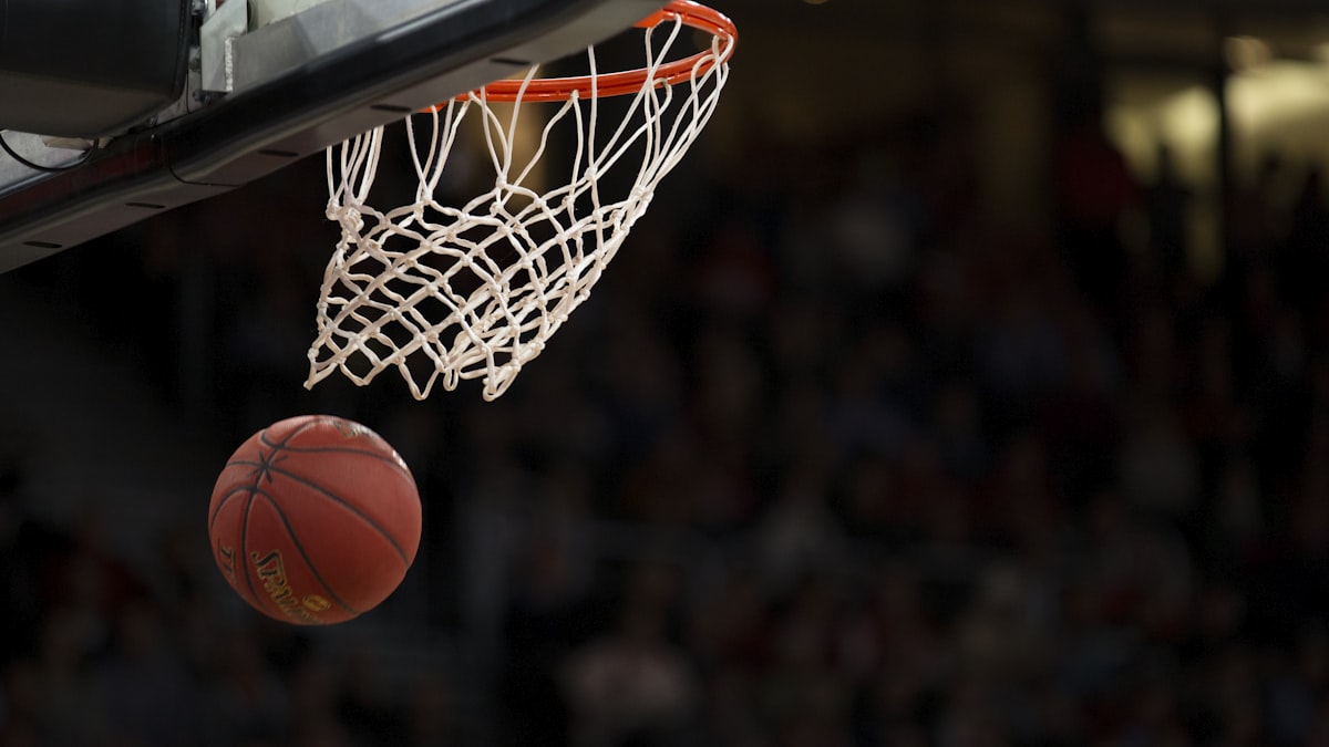 NBA篮球比赛瞬间，球员高高跃起在篮筐上方准备大力扣篮，防守球员试图封盖
